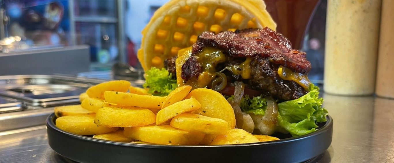 Waffel-Cheeseburger & Select Crisp Fries Burger mit Waffelbrötchen, Salat, Bacon und frittierten Bratkartoffeln auf einem schwarzen Teller.